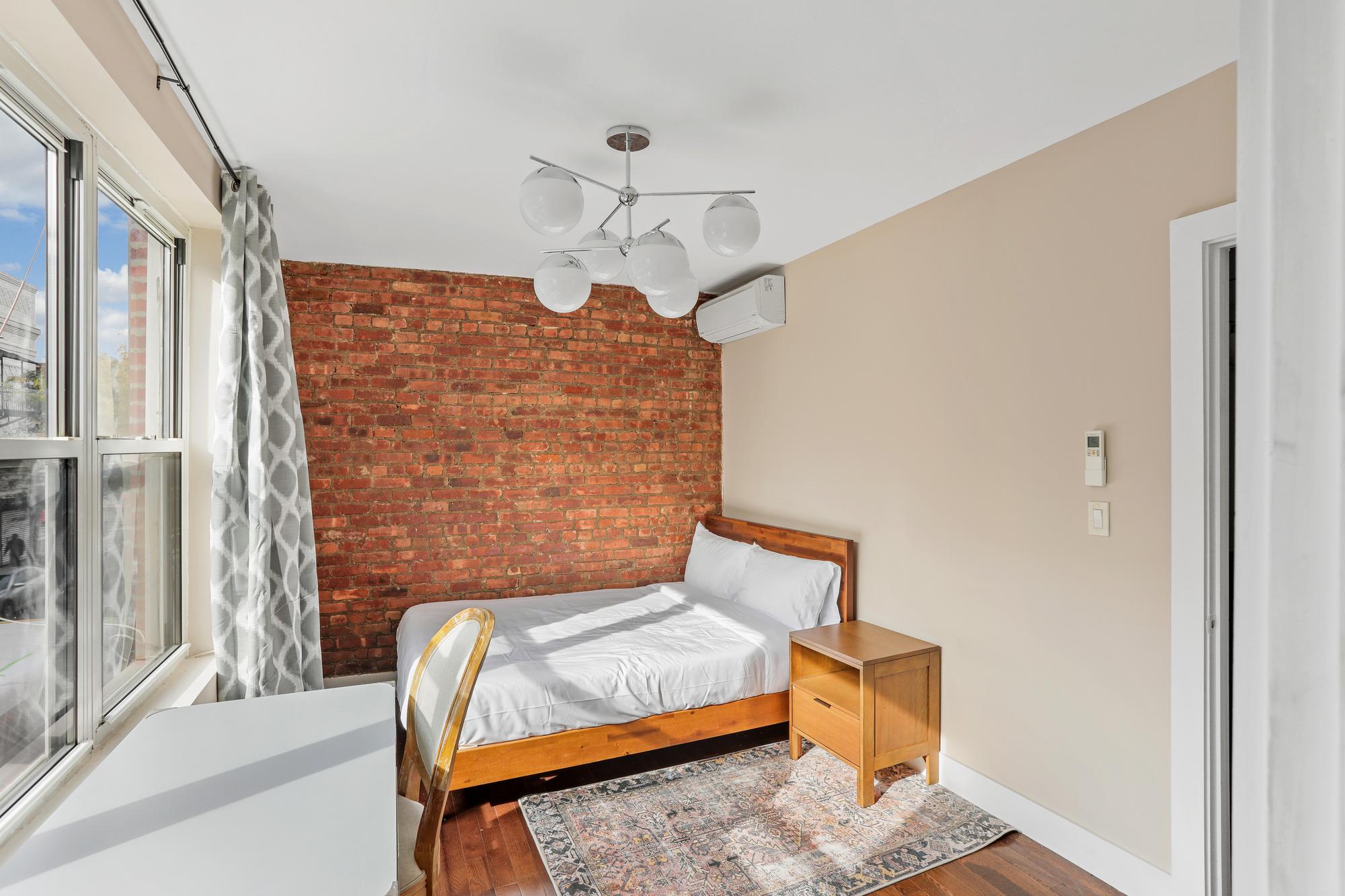 A cozy bedroom with a large window, brick accent wall, and modern light fixture. The room features a comfortable bed with white linens, a wooden nightstand, a wooden chair, and a patterned rug.