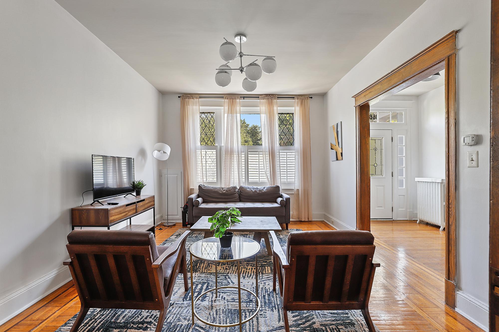 A cozy and bright living room with a mid-century modern design. The room features a comfortable grey sofa placed against a wall with large windows draped with sheer curtains, allowing plenty of natural light to flood the space. Two wooden armchairs with brown cushions face the sofa, accompanied by a round glass coffee table in the center. A contemporary light fixture with multiple round bulbs hangs from the ceiling, illuminating the room. The floor is adorned with a patterned rug, and a flat-screen TV sits on a wooden console table to the left. The room has hardwood floors and white walls, with a wooden doorway leading to a bright entryway with a white front door and sidelights.